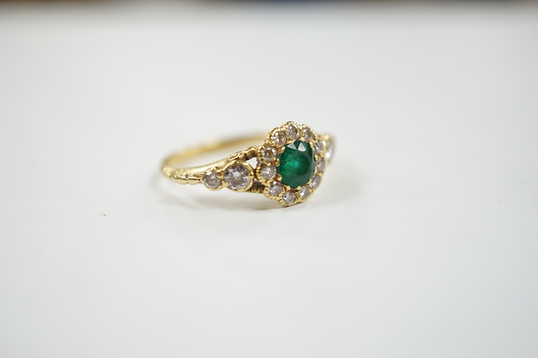 A Victorian style yellow metal emerald and diamond set circular cluster ring, with graduated diamond set shoulders, size N, gross weight 3.7 grams.